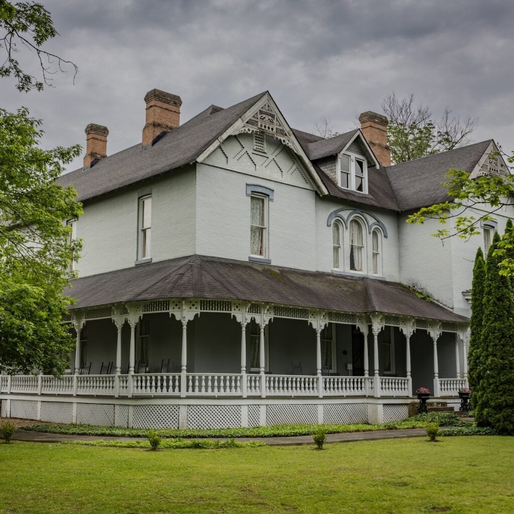 A majestic Victorian mansion painted in soft grey with intricate white gingerbread trim and a sweeping wraparound porch. The historic estate features tall brick chimneys and gabled roofs, nestled among lush green lawns and towering trees under a moody, overcast sky.