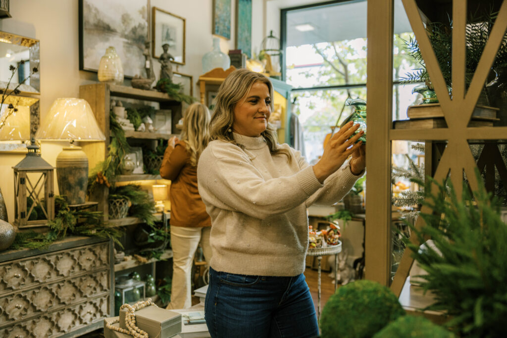 A bright and layered interior scene of a high-end boutique in McMinnville, TN, where a woman browses a shelf of elegant green glassware, surrounded by sophisticated home decor, lamps, and seasonal greenery.