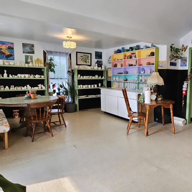 A bright and whimsical pottery studio and café interior. Rainbow-colored shelves are lined with ceramic pieces waiting to be painted, while rustic wooden tables and mismatched chairs sit under soft lighting, surrounded by vibrant local artwork and lush houseplants.