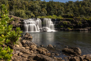 A wide, 30-foot horseshoe-shaped waterfall cascading over jagged limestone ledges into a deep river gorge, with the historic brick 19th-century cotton mill perched on the bluff above.
