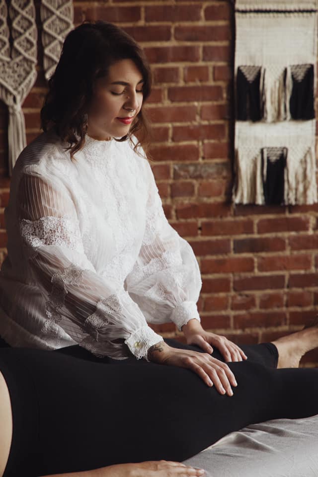 A serene wellness practitioner with dark hair and a white lace blouse performs a gentle, hands-on somatic therapy session. She sits with closed eyes in a state of quiet focus, her hands resting on a client's leg against a warm backdrop of an exposed red brick wall adorned with woven macramé tapestries.