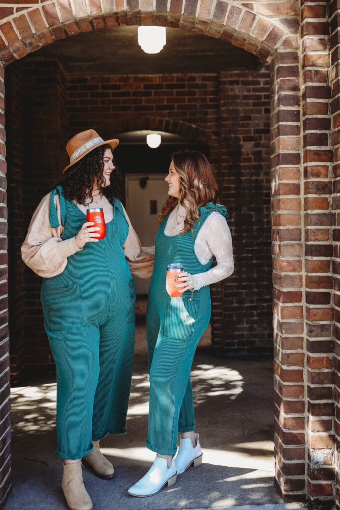 Two women wearing stylish, deep-teal overalls and hats stand laughing under a historic brick archway, holding colorful insulated tumblers.