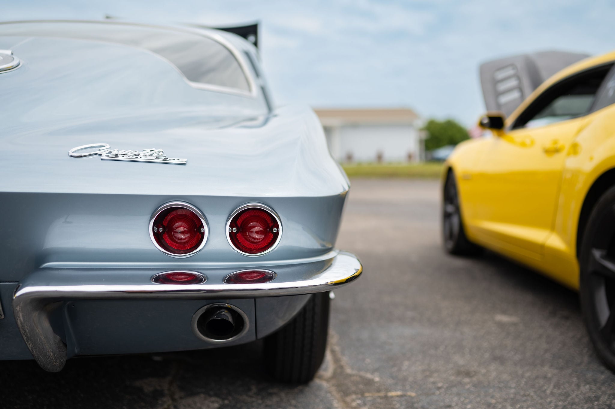 A close-up, low-angle rear view of a vintage light blue Corvette Stingray showing its iconic round dual taillights and chrome bumper, with a modern yellow sports car blurred in the background.