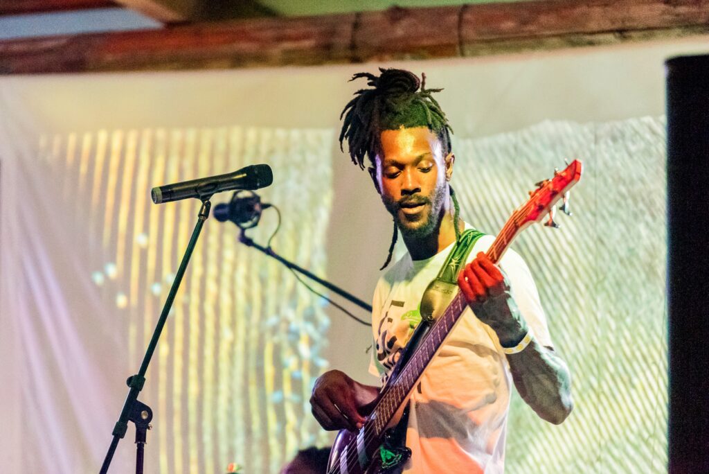 A male bassist with long dreadlocks tied up performs on stage, illuminated by a warm spotlight against a backdrop of glowing vertical light patterns.