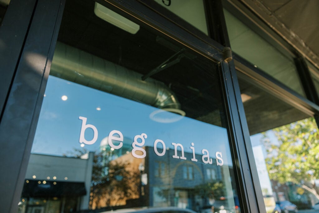 A close-up shot of a glass storefront window with the word "begonias" written in elegant, lowercase white lettering. Reflections of a clear blue sky, green trees, and historic downtown buildings dance across the glass, while industrial ductwork is visible inside the modern restaurant space.