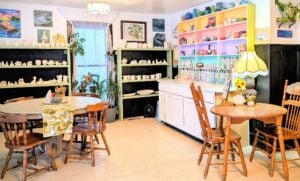 A bright and whimsical pottery studio and café interior. Rainbow-colored shelves are lined with ceramic pieces waiting to be painted, while rustic wooden tables and mismatched chairs sit under soft lighting, surrounded by vibrant local artwork and lush houseplants.
