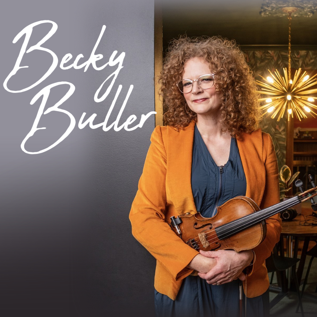 A portrait of bluegrass artist Becky Buller with curly hair and glasses, wearing a vibrant orange blazer and holding a wooden fiddle against her chest in a room with a modern gold starburst chandelier.