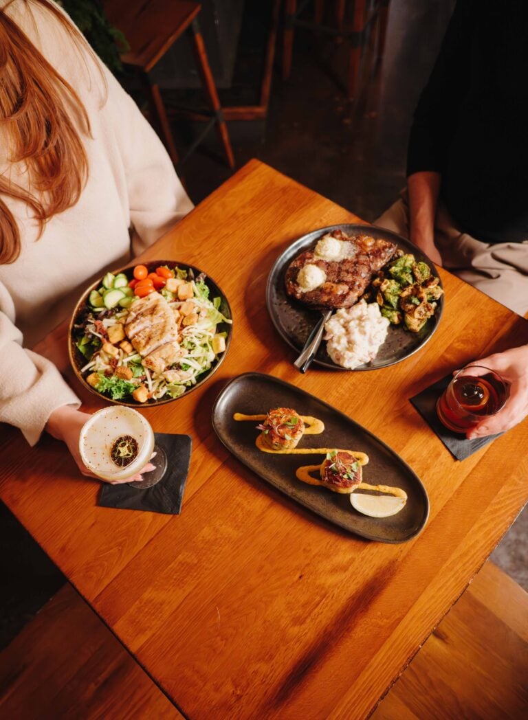 An overhead view of a dinner spread at The Local on Morford, featuring a grilled steak with butter, a fresh chicken salad, seared scallops, and craft cocktails.