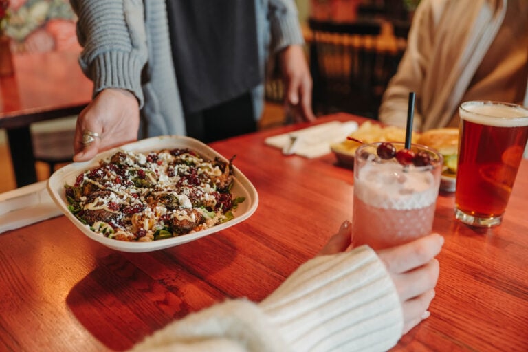 A server presenting a fresh bowl of roasted brussels sprouts with goat cheese at Barren Fork Brews alongside a craft cocktail and a pint of beer.