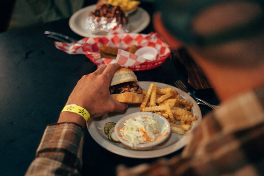 A couple sits at a rustic wooden table in a local BBQ joint, smiling as they share a platter of pulled pork, ribs, and classic sides like mac and cheese and cornbread.