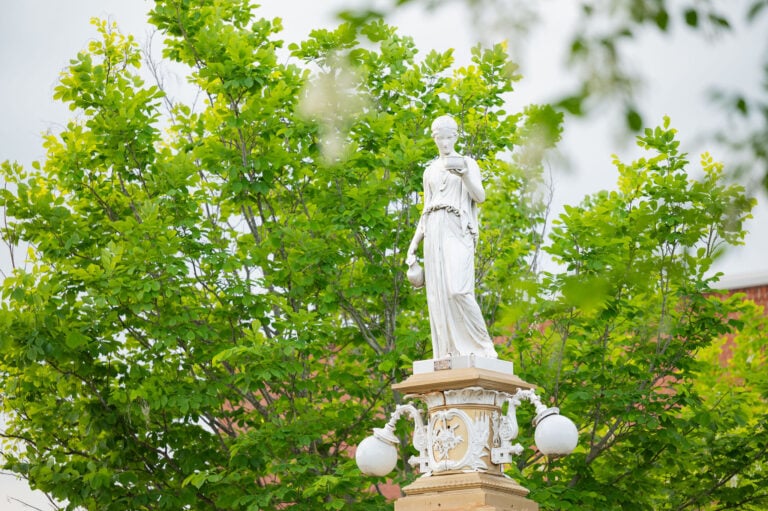 A classical white statue of the goddess Hebe standing atop an ornate pedestal surrounded by lush green trees.