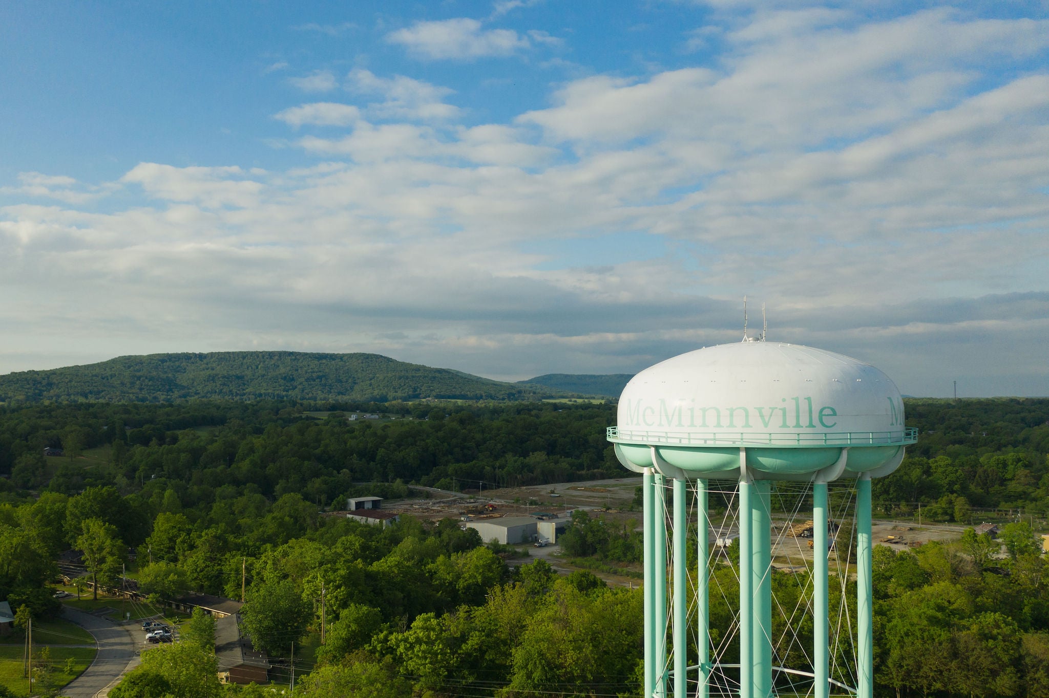 An aerial view of the mint-green McMinnville water tower overlooking a lush, forested landscape and the rolling hills of the Cumberland Plateau.
