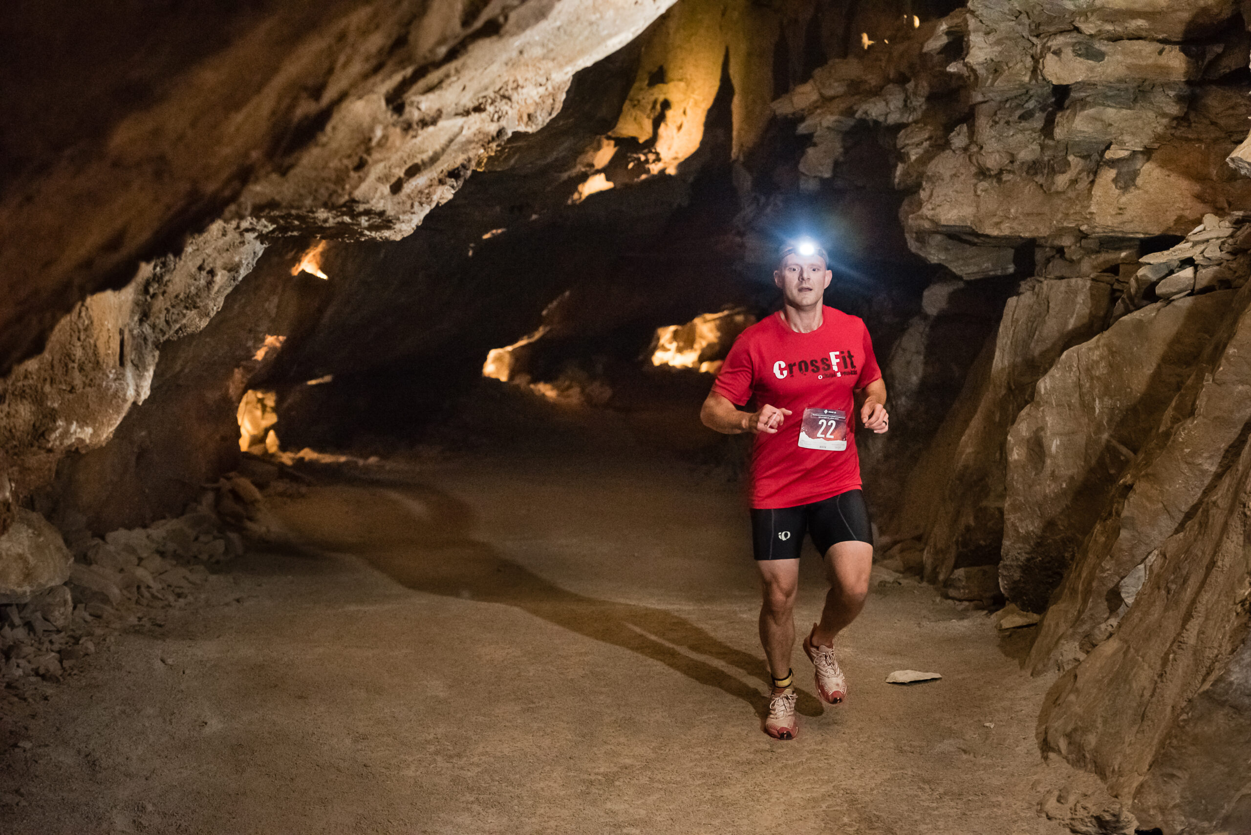 A male runner in a red shirt and headlamp navigates a wide, sandy-floored passage deep within Cumberland Caverns, surrounded by rugged limestone walls and warm, ambient cave lighting.