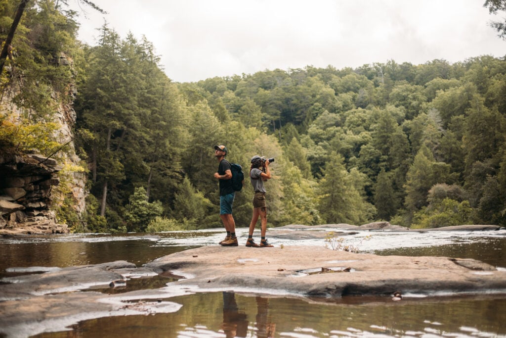 Two hikers stand back-to-back on a flat rock in the middle of a shallow river at the top of a waterfall, surrounded by dense green forest and towering limestone cliffs under an overcast sky.