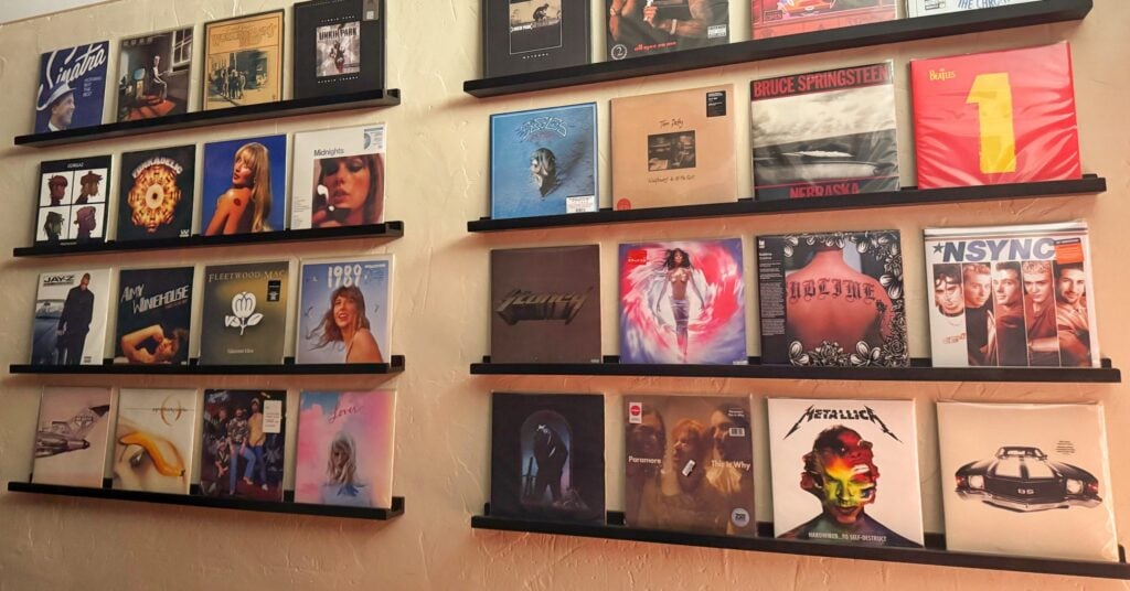 A gallery wall of vinyl record albums, including Taylor Swift and Metallica, displayed on black floating shelves against a warm, textured cream wall.