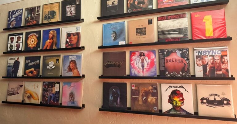 A gallery wall of vinyl record albums, including Taylor Swift and Metallica, displayed on black floating shelves against a warm, textured cream wall.