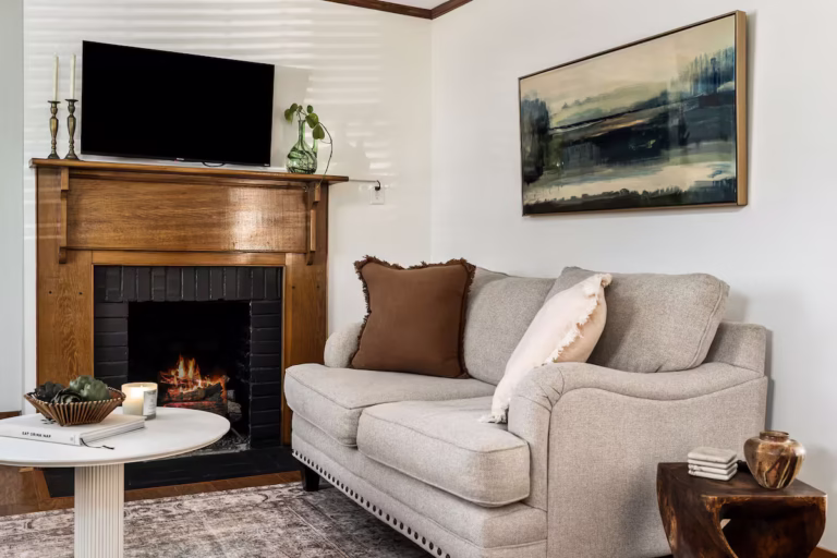 A wide-angle interior view of a spacious, light-filled living room featuring a mix of mid-century and modern furniture, with large windows overlooking a historic McMinnville street.