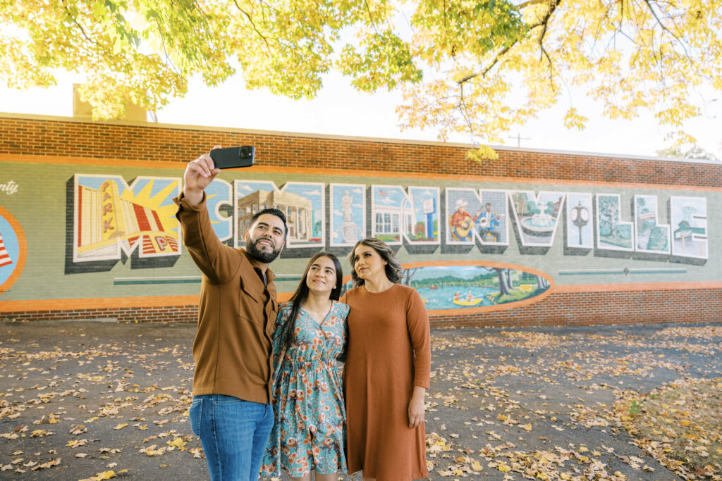 A vibrant, large-scale outdoor mural stretching across a city building, featuring bold colors and intricate artistic details. A small group of travelers stands nearby, taking a selfie with the artwork under a clear sky.