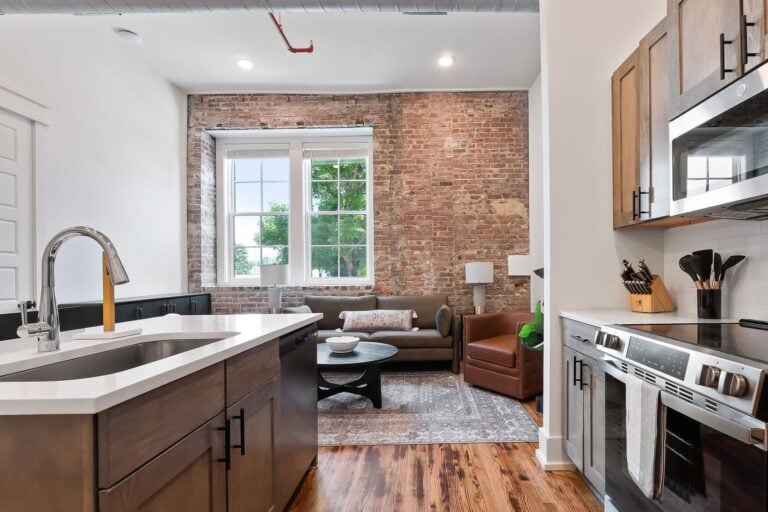 An airy open-concept apartment featuring an exposed brick wall, warm hardwood floors, and a modern kitchen. A soft olive-green sofa and a leather armchair sit by a large window that floods the living space with natural light.