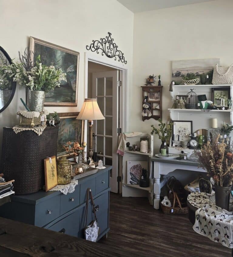 The interior of a boutique shop featuring a large crystal chandelier, vintage blue dressers, framed landscape art, and floral arrangements.
