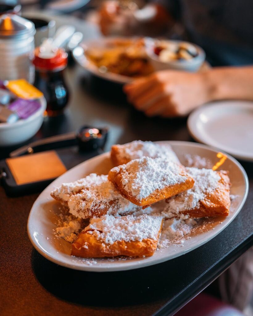 A close-up of a white oval plate piled high with golden-brown, square beignets generously dusted with a thick layer of powdered sugar, set on a dark restaurant table.