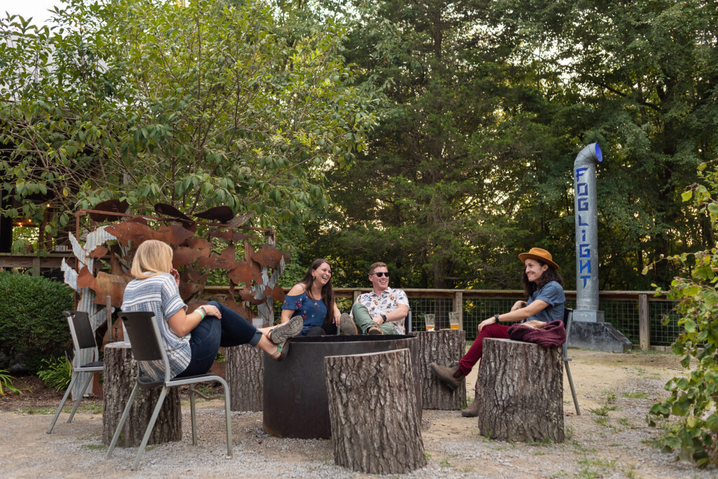 A rustic, tin-roofed restaurant tucked into a lush forest overlook, featuring a warm glow from interior lights and an eclectic porch adorned with lanterns and strings of lights above the Caney Fork River.