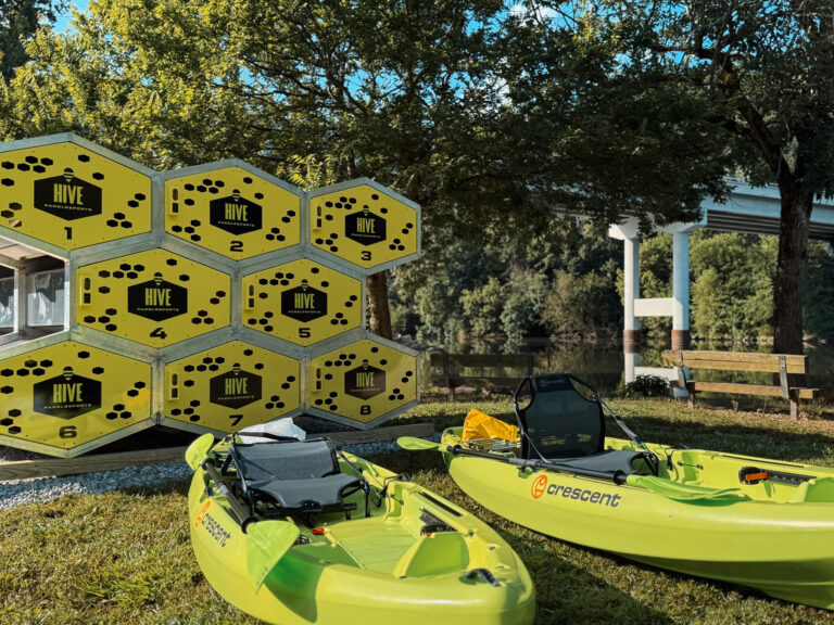 A modern, self-service kayak rental kiosk with a distinctive hexagonal "beehive" design, located at Riverfront Park alongside the scenic Barren Fork River.
