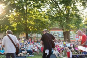 A live band performs on an outdoor stage at sunset for a large crowd seated in lawn chairs on the McMinnville town square.