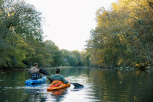 A couple in colorful kayaks paddling on a calm river surrounded by vibrant autumn trees.
