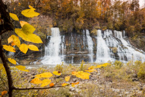 A massive 80-foot waterfall cascading out of a rocky cliff face into a river gorge surrounded by autumn trees.