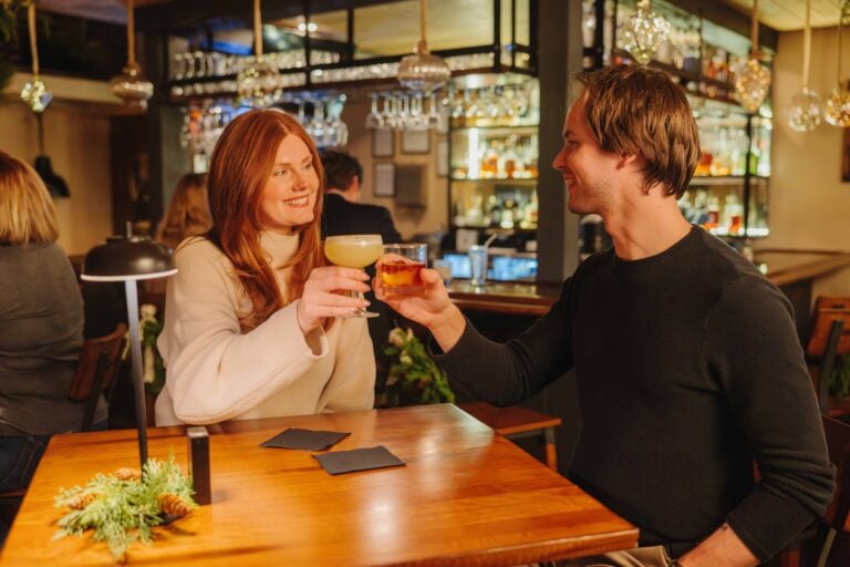 A couple toasting with craft cocktails at a polished wooden table in the warm, atmospheric interior of The Local on Morford.