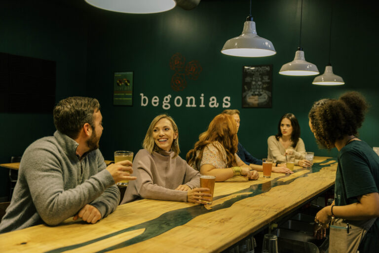 A group of friends laughing and enjoying drinks at the custom wood-and-resin river table inside the dark green dining room of Begonia's.
