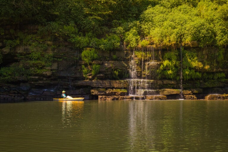 A yellow kayak gliding past cascading waterfalls and towering limestone bluffs on the Caney Fork River in Rock Island, Tennessee.