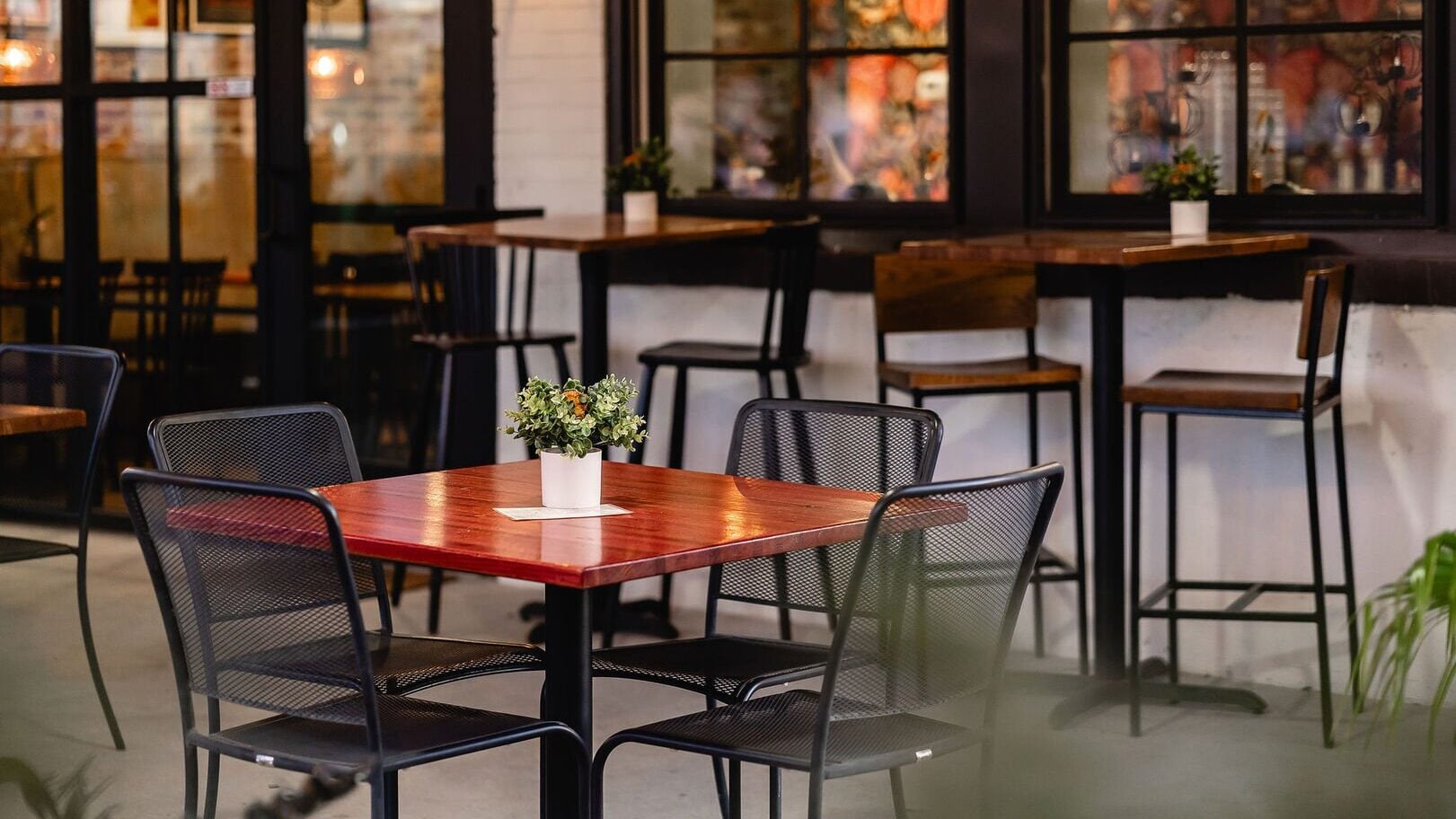 The outdoor patio of Barren Fork Brews featuring rich wooden tables and black mesh bistro chairs. Warm amber light glows through large windowpanes, casting a cozy reflection over the inviting seating area.